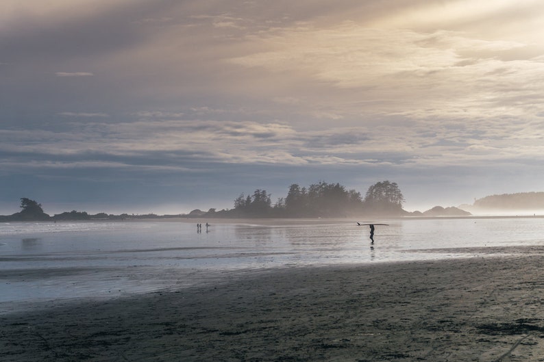 Puede incluir: Una figura solitaria camina por una playa de arena llevando una tabla de surf, con una peque&ntilde;a isla en la distancia y un cielo nublado arriba.