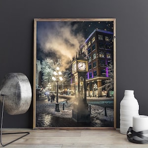 May include: A framed photograph of a snowy street scene at night. A tall clock tower with a lit clock face is in the center. Steam rises from the top of the tower. Buildings and streetlights line the street. The image is framed in a wooden frame.