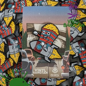 May include: A red and white striped popcorn bucket character wearing a blue and white rocket with yellow flames. The character is smiling and has large eyes. The rocket is flying through the air. The background is a sunset scene with a drive-in movie theater. The text "Vinyl Bangtan" and "Film Impact Georgia" is visible on the image.