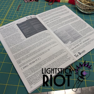 Puede incluir: Libro abierto con texto, titulado "The Diaries", sobre una esterilla de corte verde. Las p&aacute;ginas muestran texto detallado y un gr&aacute;fico. El logotipo "LIGHTSTICK RIOT" es visible en la parte inferior derecha, con una peque&ntilde;a ilustraci&oacute;n.