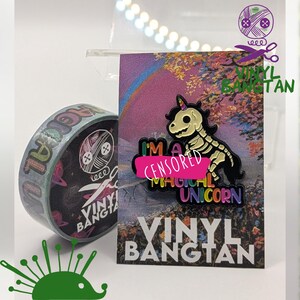 May include: A colorful enamel pin featuring a skeleton unicorn with rainbow text that reads "I'm a magical unicorn". The pin is on a card with a rainbow background and the text "Vinyl Bangtan".
