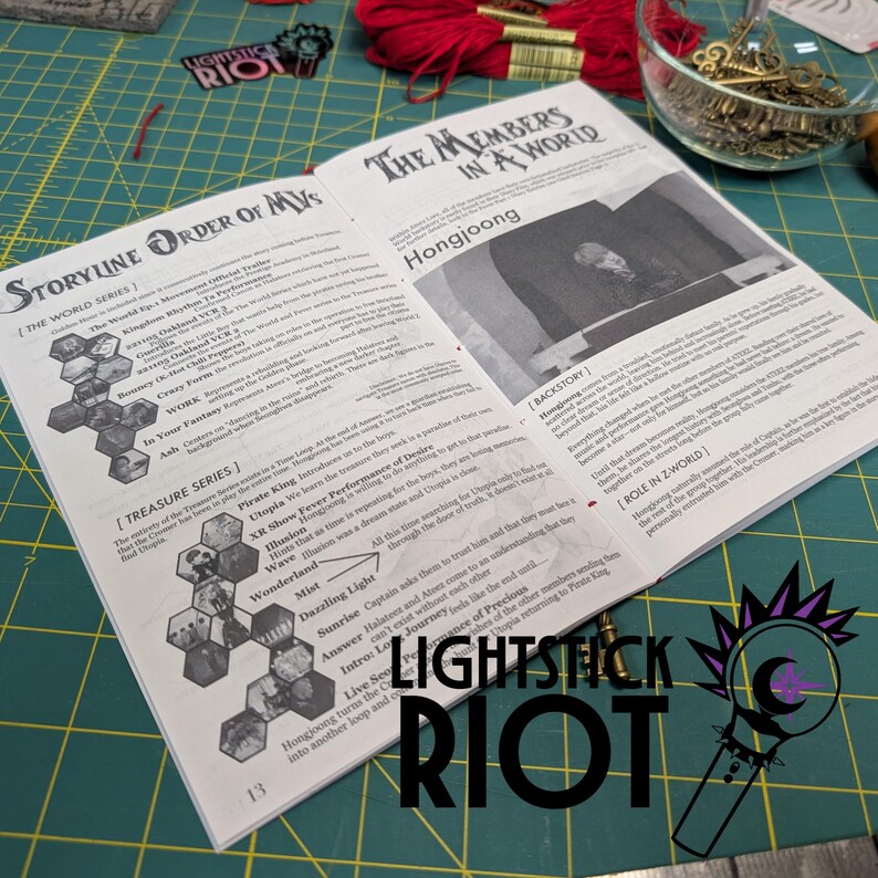 Puede incluir: Libro abierto con texto sobre una serie de historias y tesoros, con el texto "LIGHTSTICK RIOT" en la parte inferior. El libro est&aacute; sobre una esterilla de corte verde, con hilo rojo y llaves en el fondo.