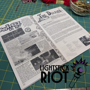 Puede incluir: Peri&oacute;dico abierto con los titulares "ANITEEZ THE LORE" y "THE DIARIES". Las p&aacute;ginas est&aacute;n llenas de texto e ilustraciones. La esquina inferior derecha tiene el texto "LIGHTSTICK RIOT" y un logotipo. El peri&oacute;dico est&aacute; sobre una esterilla de corte verde.