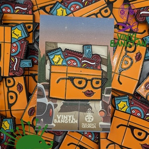 May include: A close-up of a stack of orange enamel pins featuring a cartoon character with glasses and a red mouth. The character is holding a box filled with colorful snacks. The pins are labeled "Vinyl Bangtan" and "Film Impact Georgia".