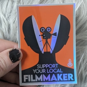 May include: A holographic sticker with an orange background and a black silhouette of a person with a film camera inside their head. The text reads "Support Your Local Filmmaker".