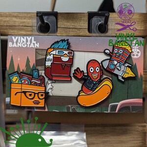 May include: Four enamel pins featuring cartoon characters. The pins are on a card with the text "Vinyl Bangtan". The characters are a red and white striped character riding a rocket, a red character with a hot dog body riding a hot dog, a red and brown character with a 3D glasses on its head, and a yellow character with glasses and a box of snacks.