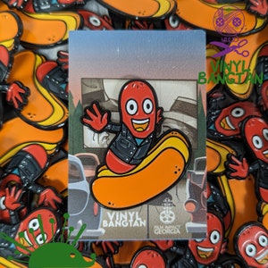 May include: A red and black enamel pin featuring a cartoon hot dog wearing a leather jacket and riding a hot dog shaped skateboard. The pin says "Vinyl Bangtan" and "Film Impact Georgia".