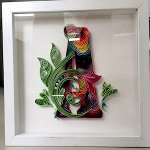 Op de afbeelding: Een ingelijst papieren kunstwerk van de letter A met een regenboogkleurenschema en groene bladeren. De letter is gemaakt van gekruld papier en is omgeven door een wit frame.