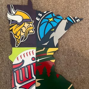 May include: A wooden outline of the state of Minnesota, featuring four different sports team logos. The top left is a purple and gold Minnesota Vikings logo. The top right is a blue and grey Minnesota Wild logo. The bottom left is a white, red and blue Minnesota Twins logo. The bottom right is a green and red Minnesota Timberwolves logo.