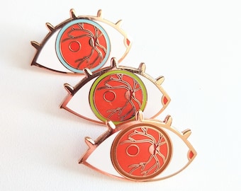 May include: Three enamel pins shaped like eyes with white outlines and red interiors. The eyes have a detailed anatomical design of the optic nerve and blood vessels in gold.