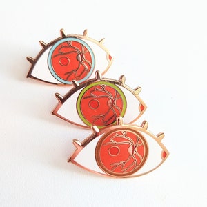 May include: Three enamel pins shaped like eyes with white outlines and red interiors. The eyes have a detailed anatomical design of the optic nerve and blood vessels in gold.