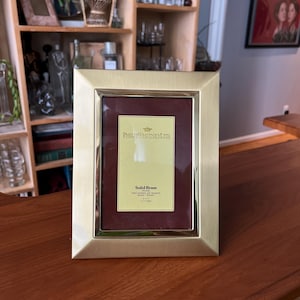 May include: A gold-coloured brass picture frame with a burgundy mount. The frame has a simple, elegant design. The text "PHILIP WHITNEY LTD" is visible, along with "Solid Brass" and dimensions of 12.7cm x 17.8cm. The frame is displayed on a wooden surface.