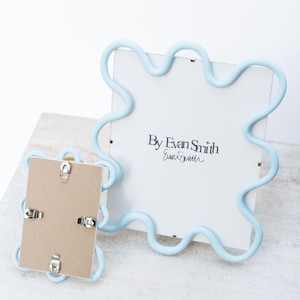 May include: Two light blue wavy-edged picture frames. The larger frame displays the text By Evan Smith with a signature below. The smaller frame is shown from the back, revealing a brown cardboard backing secured with four silver metal clips.