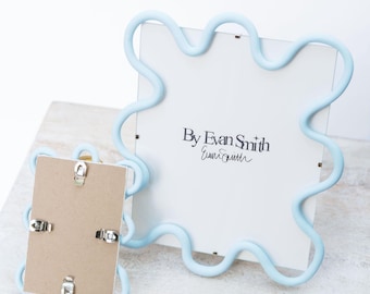 May include: Two light blue wavy-edged picture frames. The larger frame displays a white insert with the text By Evan Smith. The smaller frame is positioned in front, showing its brown cardboard backing with four silver metal swivel clips.