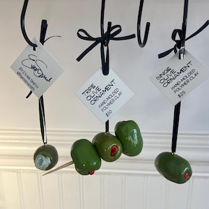 Könnte beinhalten: Handgeformte Polymer-Ton-Oliven-Ornamente, die an schwarzen Haken hängen. Einige haben rote Akzente, andere einen Zahnstocher. Auf den Etiketten steht "Tripe Olive Ornament" und "Single Olive Ornament".