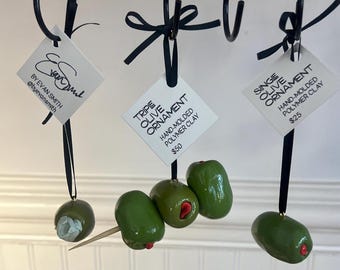 Può includere: Ornamenti a forma di oliva in argilla polimerica modellata a mano appesi a ganci neri. Alcuni hanno accenti rossi, altri uno stuzzicadenti. Le etichette recitano "Tripe Olive Ornament" e "Single Olive Ornament".
