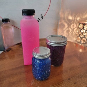 May include: Three glass jars with glitter inside. The jars have silver lids. The jar on the left is filled with blue glitter, the jar in the middle is filled with purple glitter, and the jar on the right is filled with pink glitter.