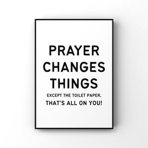 Op de afbeelding: Een ingelijste witte print met zwarte tekst: "PRAYER CHANGES THINGS EXCEPT THE TOILET PAPER, THAT'S ALL ON YOU!" De print heeft een zwarte rand en wordt weergegeven tegen een witte achtergrond.