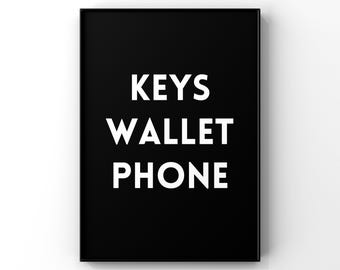 May include: A rectangular wall art print featuring a solid black background with the white sans-serif text KEYS, WALLET, and PHONE stacked vertically in the center. The print is enclosed within a thin black frame, creating a high-contrast design.