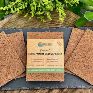K&ouml;nnte beinhalten: Vier braune Kokosnuss-Schrubbpads auf einer schwarzen Schieferoberfl&auml;che. Die Schrubbpads bestehen aus nat&uuml;rlichen Fasern und sind f&uuml;r die Reinigung von Geschirr und anderen Oberfl&auml;chen konzipiert. Die Verpackung der Schrubbpads ist auf dem Bild sichtbar und enth&auml;lt den Text "MARASOL Coconut SCRUB PAD and BAR SOAP SAVER Tough Cleaner Non-Scratch Keeps Soap Dry No Gooey Mess Odorless - Antibacterial Easy Rinse - Quick Dry ALL NATURAL PLASTIC FREE | COMPOSTABLE".