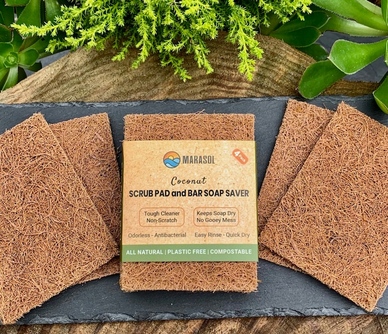 K&ouml;nnte beinhalten: Vier braune Kokosnuss-Schrubbpads auf einer schwarzen Schieferoberfl&auml;che. Die Schrubbpads bestehen aus nat&uuml;rlichen Fasern und sind f&uuml;r die Reinigung von Geschirr und anderen Oberfl&auml;chen konzipiert. Die Verpackung der Schrubbpads ist auf dem Bild sichtbar und enth&auml;lt den Text "MARASOL Coconut SCRUB PAD and BAR SOAP SAVER Tough Cleaner Non-Scratch Keeps Soap Dry No Gooey Mess Odorless - Antibacterial Easy Rinse - Quick Dry ALL NATURAL PLASTIC FREE | COMPOSTABLE".