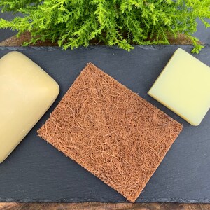 K&ouml;nnte beinhalten: Zwei Seifenst&uuml;cke, eines beige und eines gelb, liegen auf einer schwarzen Schieferfl&auml;che mit einem braunen Kokosfaser-Schwamm in der Mitte.