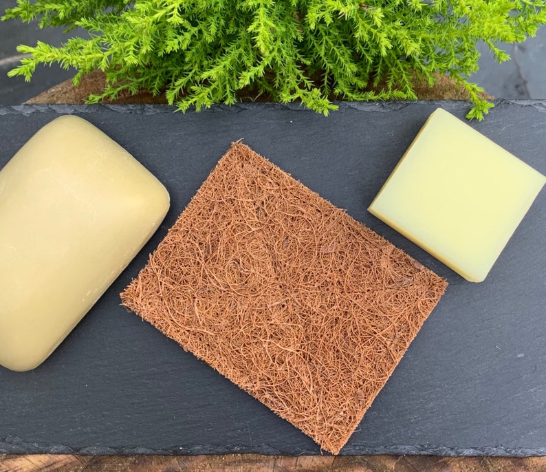 K&ouml;nnte beinhalten: Zwei Seifenst&uuml;cke, eines beige und eines gelb, liegen auf einer schwarzen Schieferfl&auml;che mit einem braunen Kokosfaser-Schwamm in der Mitte.