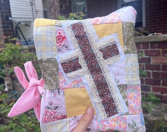 May include: A patchwork cushion with a cross design. The cushion features floral and patterned fabrics in pink, yellow, and green. The cross is made of patterned fabric with lace trim. A pink fabric bow is attached to the side.