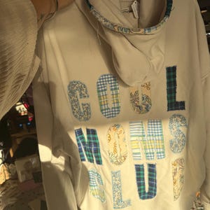 以下が含まれることがあります： さまざまな柄の生地で背中に「COOL MOMS CLUB」の文字がアップリケされたクリーム色のパーカー。フードとネックラインは花柄の生地でトリミングされています。パーカーは木製のハンガーに吊るされています。