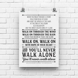 Könnte beinhalten: Ein gerahmter Druck mit dem Text von "You'll Never Walk Alone" in schwarzer Schrift auf cremefarbenem Hintergrund. Der Druck wird von silbernen Clips an einer weißen Backsteinmauer gehalten. Der Text enthält den Titel und den Künstler des Liedes.