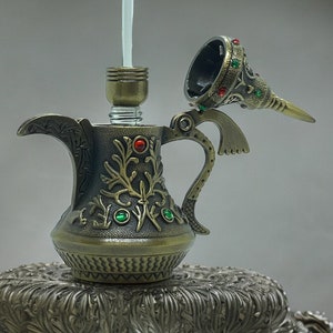 以下が含まれることがあります： ブロンズ仕上げの、装飾的なアンティークスタイルの水タバコ。赤い宝石と緑の宝石のアクセントが付いた装飾的な花の模様が特徴です。透明なガラス管と詳細なマウスピースも確認できます。