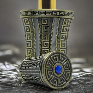Op de afbeelding: Een decoratieve parfumfles met een goudkleurige sproeikop en een gedetailleerd ontwerp in antieke stijl. De fles heeft een herhalend geometrisch patroon in goud en donkergrijs, en de dop heeft een blauwe edelsteen.