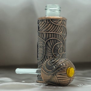 以下が含まれることがあります： 装飾的なデザインの、ブロンズ色のガラス瓶。透明なガラスのトップと、黄色のアクセントが付いたお揃いの球形のストッパーが付いています。デザインは渦巻き模様が特徴です。