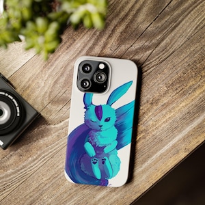 May include: A white phone case with a blue and purple cartoon bunny illustration. The bunny has a white face with a purple nose and blue eyes. The bunny is curled up in a ball with its tail wrapped around its body.