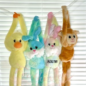 May include: Four plush hanging toys, a yellow duck, a blue and green bunny, a pink and white bunny, and a brown monkey. The pink and white bunny has the name "Jazalynn" written on it.