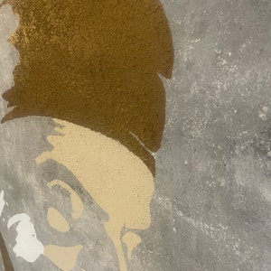 May include: A close-up of a gold and beige painted stencil of a person's face on a gray textured background.