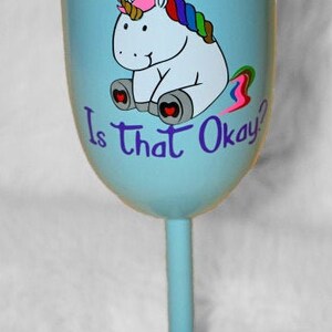 May include: A light blue wine glass with a unicorn design and the text "Is that Okay?" The unicorn is white with a rainbow mane and tail. The glass has a gold lid.