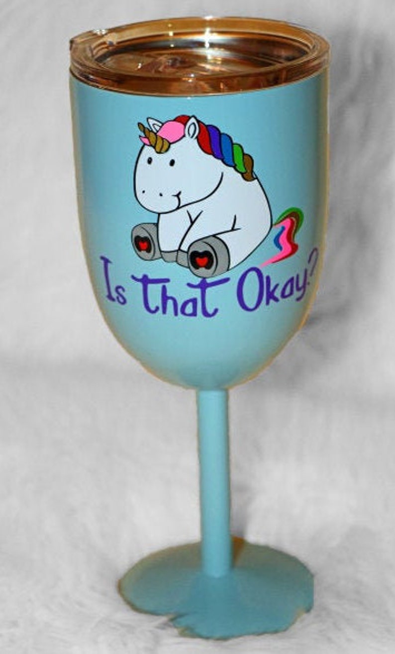May include: A light blue wine glass with a unicorn design and the text "Is that Okay?" The unicorn is white with a rainbow mane and tail. The glass has a gold lid.