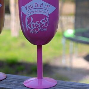 May include: Pink insulated wine glass with a white graduation cap design and the text "You Did It! Congratulations Rossy Wife Mom".