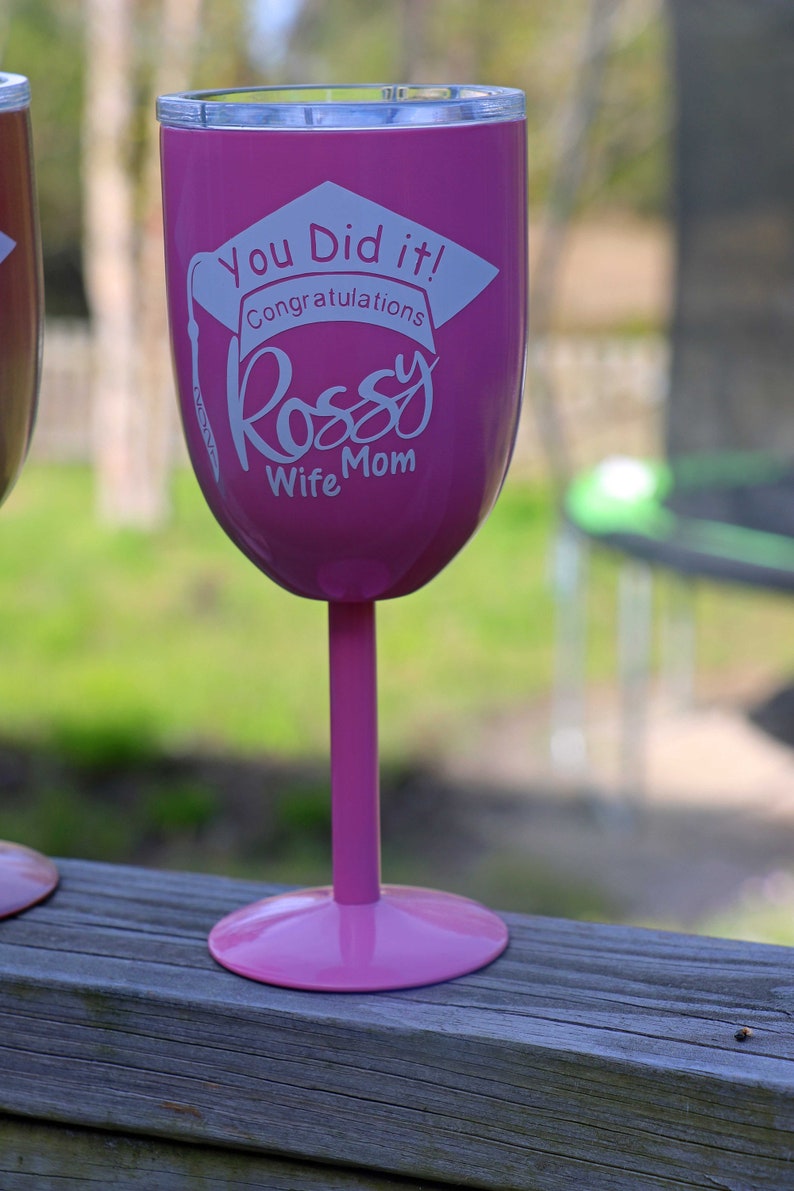 May include: Pink insulated wine glass with a white graduation cap design and the text "You Did It! Congratulations Rossy Wife Mom".