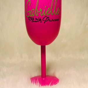 May include: A pink wine glass with a lid. The glass has the name "Gabrielle" written in green cursive lettering and the words "Fresh Princess" written in black lettering below the name.