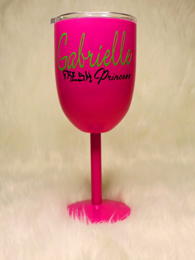 May include: A pink wine glass with a lid. The glass has the name "Gabrielle" written in green cursive lettering and the words "Fresh Princess" written in black lettering below the name.