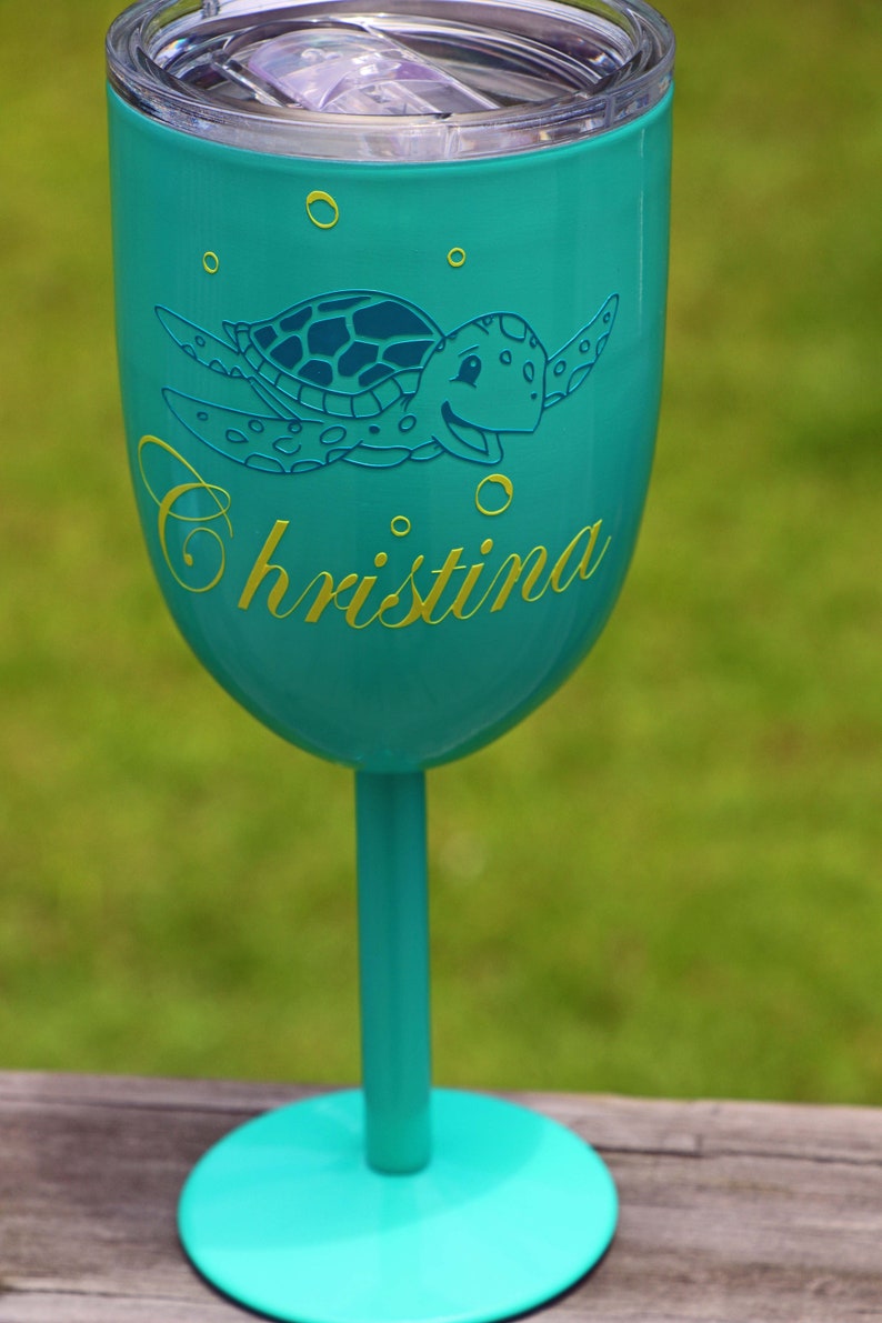 May include: A turquoise blue stainless steel wine tumbler with a lid and a sea turtle design. The tumbler has the name "Christina" written on it in yellow.