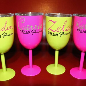 May include: Four neon-colored wine glasses with black lettering. The glasses are yellow and pink. The text on the glasses reads "London Fresh Princess", "Carrie Fresh Princess", "Zela Fresh Princess", and "Kalyiah Fresh Princess".