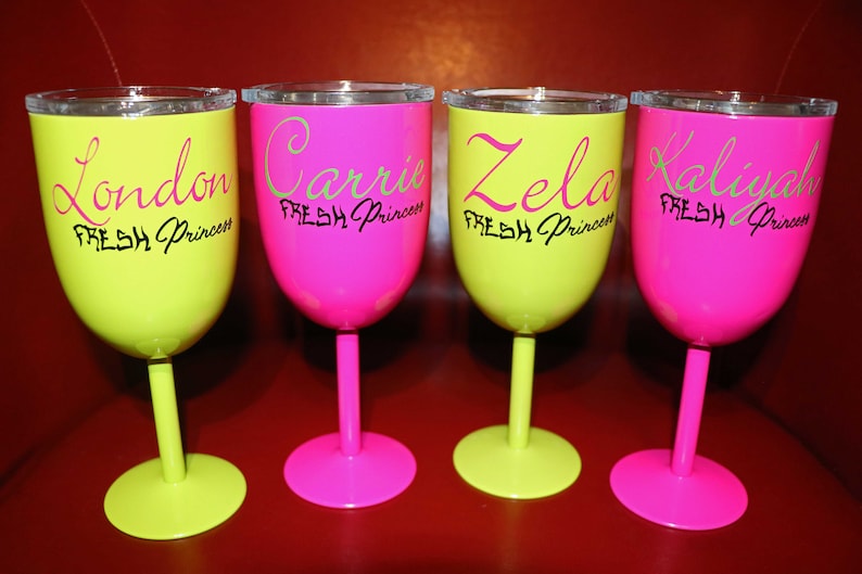 May include: Four neon-colored wine glasses with black lettering. The glasses are yellow and pink. The text on the glasses reads "London Fresh Princess", "Carrie Fresh Princess", "Zela Fresh Princess", and "Kalyiah Fresh Princess".