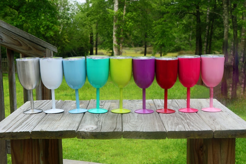 May include: Ten stainless steel wine tumblers with lids in various colors, including silver, white, light blue, turquoise, green, yellow, purple, red, pink, and light pink. The tumblers are lined up on a wooden table.