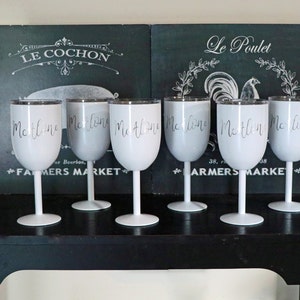 May include: Six white wine glasses with silver rims and the word "Malone" written on each glass. The glasses are on a black table in front of two black chalkboards with French words on them.