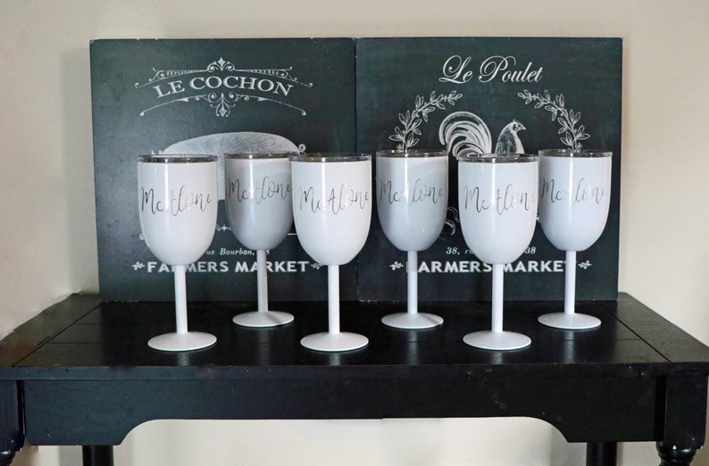 May include: Six white wine glasses with silver rims and the word "Malone" written on each glass. The glasses are on a black table in front of two black chalkboards with French words on them.
