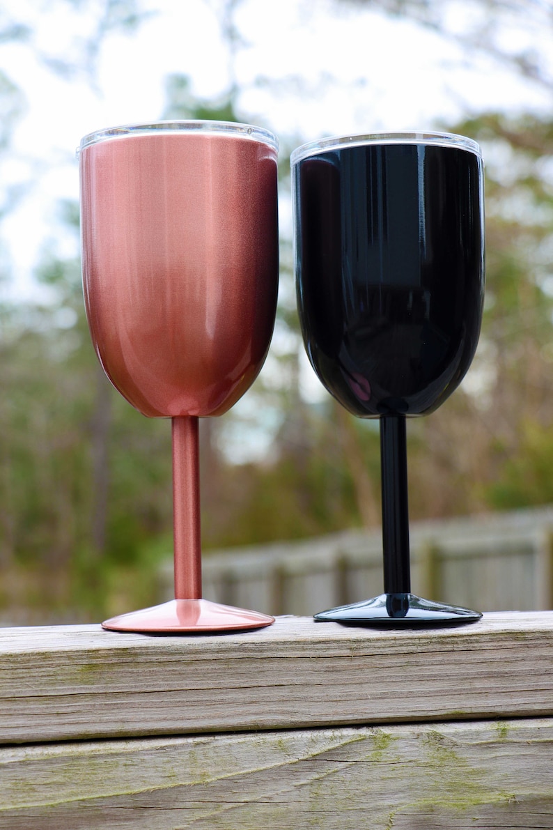 May include: Two stainless steel wine tumblers, one rose gold and one black, with a clear lid. The tumblers are on a wooden surface.