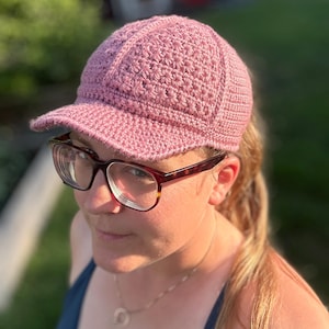 May include: A dusty rose-colored baseball cap crafted from crocheted yarn. The cap features a structured crown with a textured star-like stitch pattern and a solid brim. The design includes a multi-panel construction with visible seams.